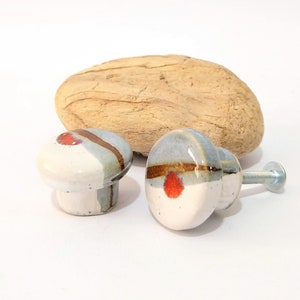 Può includere: Due pomelli per mobili in ceramica con un design bianco, grigio e rosso. Un pomello ha una vite attaccata. Un pezzo di legno marrone chiaro a forma ovale è dietro i pomelli. I pomelli sono probabilmente per mobili o decorazioni per la casa.