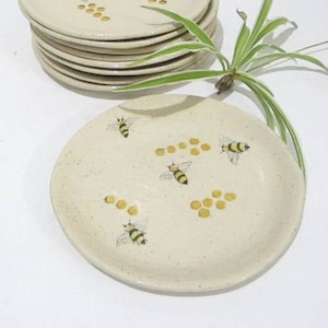 May include: A stack of six round, speckled cream-colored ceramic plates and one individual plate placed in front. Each plate features a hand-painted design of yellow and black bees and golden honeycomb patterns scattered across the surface.