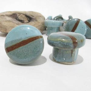 May include: A selection of ceramic cabinet knobs in blue and brown. The knobs are round with a diagonal brown stripe. A piece of driftwood is in the background. These knobs are suitable for furniture and home decor.