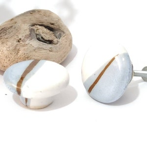 Può includere: Due pomelli per mobili in ceramica e una maniglia coordinata. I pomelli sono bianchi con una sezione diagonale azzurra e una striscia marrone. La maniglia ha un design simile. Un pezzo di legno galleggiante e una pianta verde sono sullo sfondo.