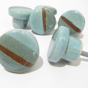 May include: A set of ceramic cabinet knobs. Each knob is round, featuring a light blue glaze and a brown stripe. The knobs have a mushroom-shaped base and a metal screw for fitting. Home decor.