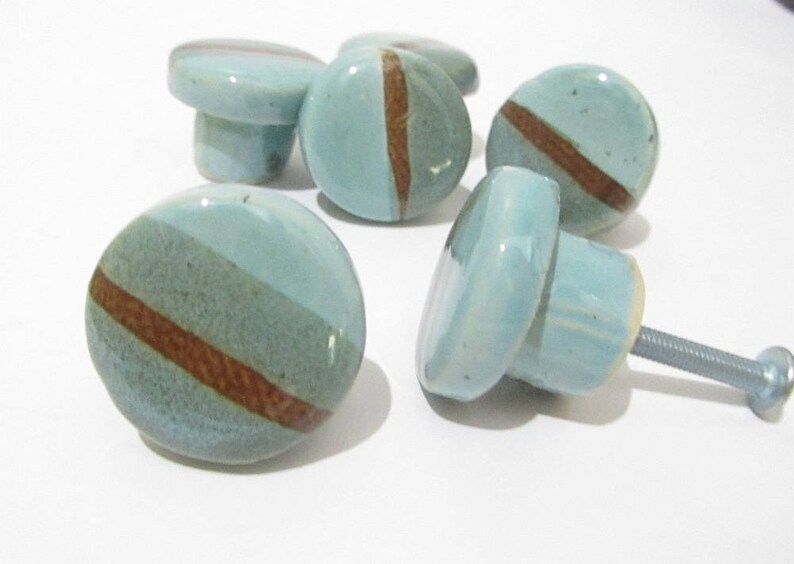 May include: A set of ceramic cabinet knobs. Each knob is round, featuring a light blue glaze and a brown stripe. The knobs have a mushroom-shaped base and a metal screw for fitting. Home decor.