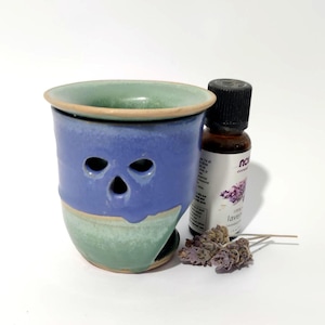 Peut inclure: Un brûle-parfum en céramique avec une partie supérieure bleue et une partie inférieure verte, doté de trois ouvertures triangulaires. À côté se trouve un petit flacon en verre ambré d'huile essentielle de lavande et un brin de lavande séchée.