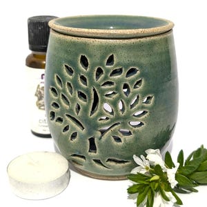 May include: A green ceramic oil warmer with a cutout tree design, featuring a speckled glaze finish. The warmer is accompanied by a small white tea light candle, a small brown glass bottle with a black cap, and a sprig of white flowers.