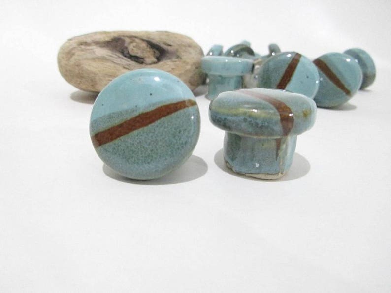 May include: A selection of ceramic cabinet knobs in blue and brown. The knobs are round with a diagonal brown stripe. A piece of driftwood is in the background. These knobs are suitable for furniture and home decor.