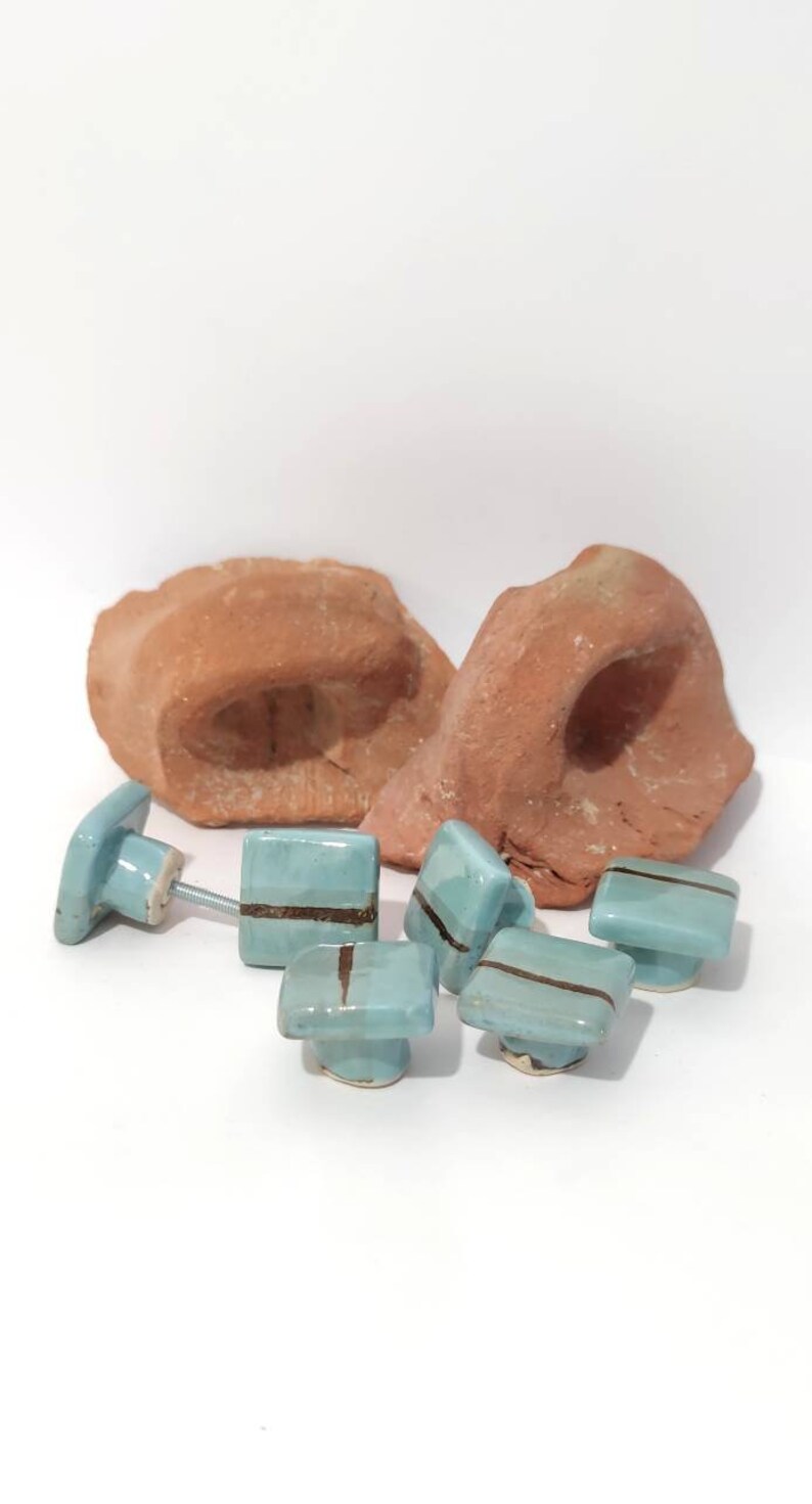 K&ouml;nnte beinhalten: Eine Sammlung hellblauer Keramik-Schrankkn&ouml;pfe mit einem dunkelbraunen Streifen, zusammen mit zwei Teilen r&ouml;tlich-brauner Keramik. Die Kn&ouml;pfe haben eine quadratische Form und sind an Metallschrauben befestigt.