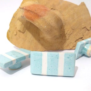 May include: Three ceramic cabinet knobs with a light blue and white striped design. The knobs are rectangular with a rounded base and a metal screw for installation. A large, textured rock is in the background.