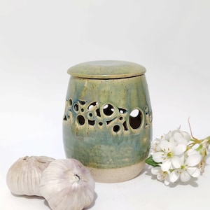 May include: A green and blue ceramic garlic keeper with a lid, decorated with circular cut-outs. Two garlic bulbs and white flowers are arranged near the keeper, against a white backdrop.