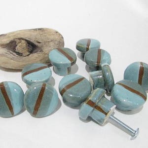 May include: A selection of ceramic cabinet knobs in teal and brown. Each knob has a teal base with a diagonal brown stripe. The knobs are round and cylindrical, with a screw for attachment. A piece of driftwood is also present.