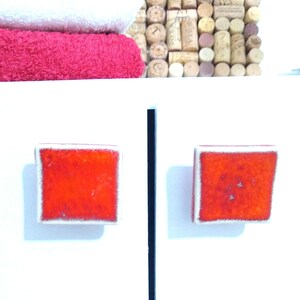 May include: Two square, red cabinet knobs with white trim are mounted on a white surface. A black vertical line is between the knobs. In the background are stacked wine corks and folded white and red towels.