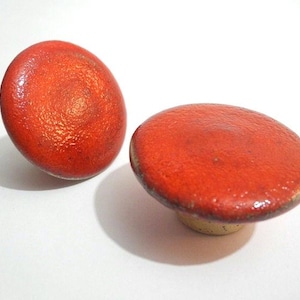 May include: Two red ceramic cabinet knobs with a textured surface. One knob is upright, the other is lying flat. The knobs have a mushroom shape and a yellow base. Home decor.