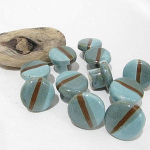 May include: A collection of ceramic cabinet knobs in shades of blue-green with a brown diagonal stripe. The knobs are round with a small handle on the back. A piece of driftwood is in the background.