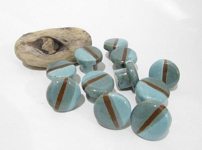 May include: A collection of ceramic cabinet knobs in shades of blue-green with a brown diagonal stripe. The knobs are round with a small handle on the back. A piece of driftwood is in the background.