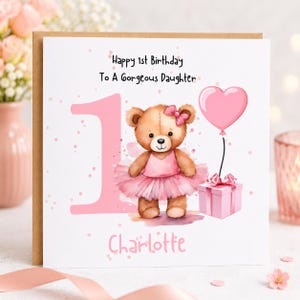 May include: A square greeting card featuring a large pink number one, a brown teddy bear wearing a pink tutu and bow, a pink heart-shaped balloon, and a pink gift box. The text reads Happy 1st Birthday To A Gorgeous Daughter and Charlotte.