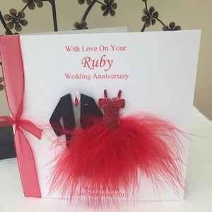 May include: A white card with red ribbon and a red feather trim. The card says "With Love On Your Ruby Wedding Anniversary". There are paper cutouts of a tuxedo and a red dress.