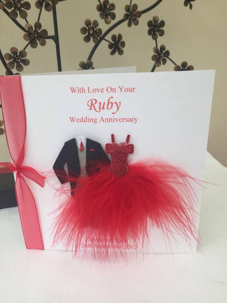 May include: A white card with red ribbon and a red feather trim. The card says "With Love On Your Ruby Wedding Anniversary". There are paper cutouts of a tuxedo and a red dress.