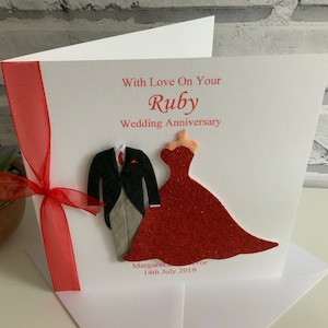 May include: A white card with red ribbon and a red glitter cutout of a bride and groom. The card says "With Love On Your Ruby Wedding Anniversary" and "Margaret & Ivor 14th July 2019".