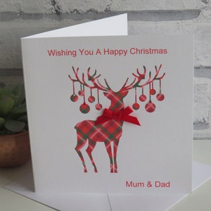 May include: A white square greeting card featuring a red and green plaid reindeer graphic with hanging ornaments on its antlers. A small red ribbon bow is attached to the reindeer's neck. The text reads Wishing You A Happy Christmas and Mum & Dad.