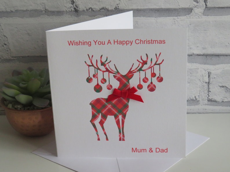 May include: A white square greeting card featuring a red and green plaid reindeer graphic with hanging ornaments on its antlers. A small red ribbon bow is attached to the reindeer's neck. The text reads Wishing You A Happy Christmas and Mum & Dad.