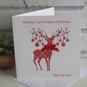 May include: A white greeting card featuring a red and green tartan reindeer illustration with hanging baubles on its antlers. A small red ribbon bow is attached to the reindeer's neck. The text reads Wishing You A Happy Christmas and Mum & Dad.