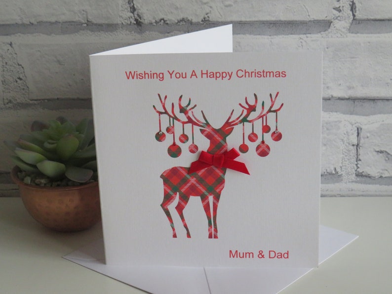 May include: A white greeting card featuring a red and green tartan reindeer illustration with hanging baubles on its antlers. A small red ribbon bow is attached to the reindeer's neck. The text reads Wishing You A Happy Christmas and Mum & Dad.