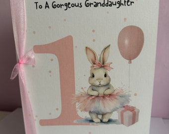 May include: A pink and white birthday card with a pink number one, a bunny wearing a pink tutu, a pink balloon, and the text "Happy 1st Birthday To A Gorgeous Granddaughter Olivia Rose".