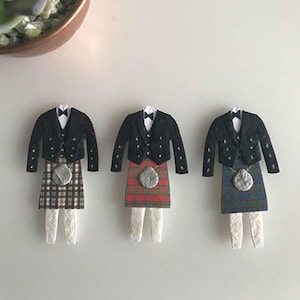 K&ouml;nnte beinhalten: Drei Papierschnitte von M&auml;nnern in traditionellen schottischen Kilts. Jeder Kilt hat ein anderes Tartanmuster: Braun und Wei&szlig;, Rot und Gr&uuml;n sowie Blau und Gr&uuml;n. Jeder Mann tr&auml;gt eine schwarze Jacke mit einem wei&szlig;en Hemd und einer schwarzen Fliege.