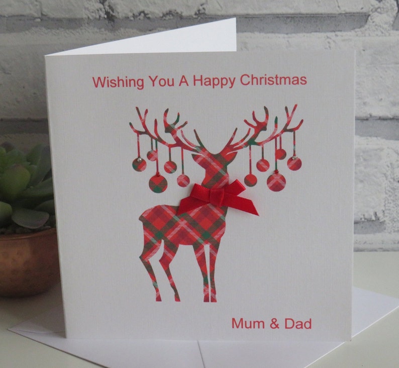 May include: A white greeting card featuring a red and green plaid reindeer silhouette with hanging ornaments on its antlers. A red ribbon bow is attached to the reindeer's neck. The text reads Wishing You A Happy Christmas and Mum & Dad.