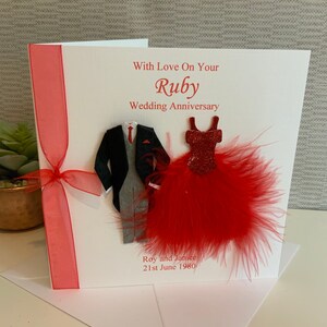 May include: A white card with a red ribbon and a red feather embellishment. The card says "With Love On Your Ruby Wedding Anniversary" and features a paper cutout of a man in a suit and a woman in a red dress. The card also says "Roy and Janice 21st June 1980".