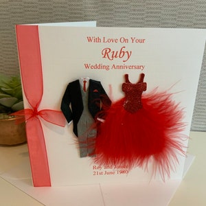 May include: A white card with a red ribbon and a red feather boa. The card says "With Love On Your Ruby Wedding Anniversary" and has a paper cutout of a man in a suit and a woman in a red dress.