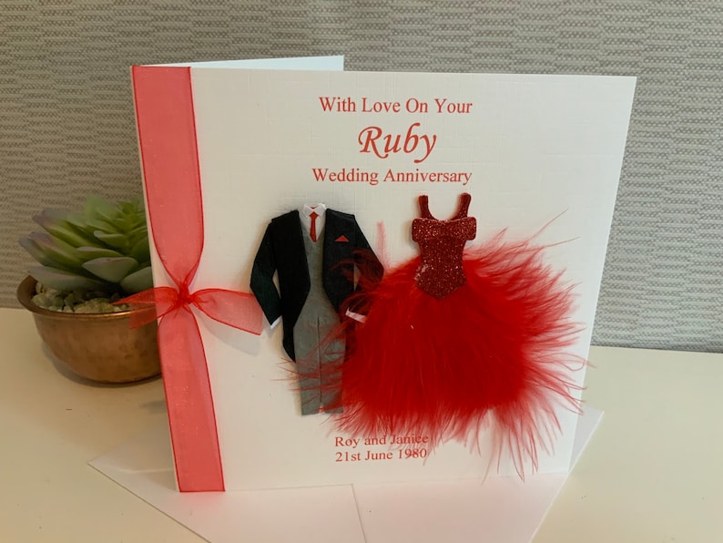 May include: A white card with a red ribbon and a red feather boa. The card says "With Love On Your Ruby Wedding Anniversary" and has a paper cutout of a man in a suit and a woman in a red dress.