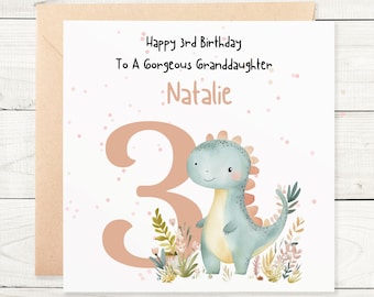 Puede incluir: Una tarjeta blanca con lunares rosas y marrones. La tarjeta dice "Happy 3rd Birthday To A Gorgeous Granddaughter: Natalie". Un gran número 3 rosa y un dinosaurio azul con barriga amarilla están en la tarjeta.