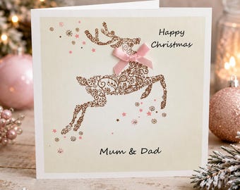 May include: A square greeting card with a cream background featuring a brown filigree reindeer illustration. The reindeer has a pink satin bow at its neck. The text reads Happy Christmas and Mum & Dad. Small pink stars and snowflakes surround the deer.