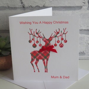 May include: A white square greeting card featuring a red and green plaid reindeer silhouette with hanging ornaments on its antlers. A red ribbon bow is attached to the reindeer's neck. The text reads Wishing You A Happy Christmas and Mum & Dad.