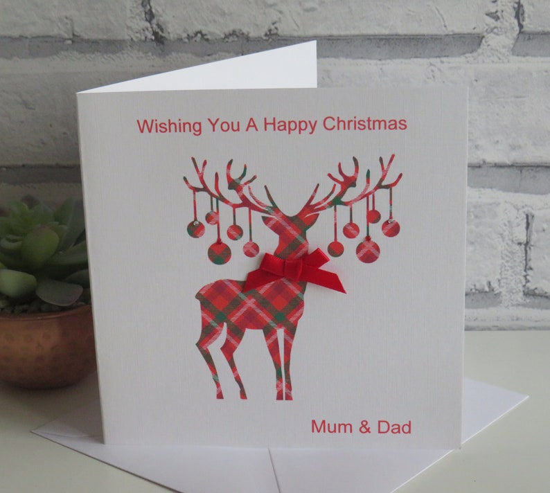 May include: A white square greeting card featuring a red and green plaid reindeer silhouette with hanging ornaments on its antlers. A red ribbon bow is attached to the reindeer's neck. The text reads Wishing You A Happy Christmas and Mum & Dad.