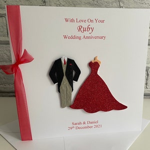 May include: A white card with red ribbon and a red glitter dress and a black suit with a grey waistcoat. The card says "With Love On Your Ruby Wedding Anniversary" and "Sarah & Daniel 29th December 2021".
