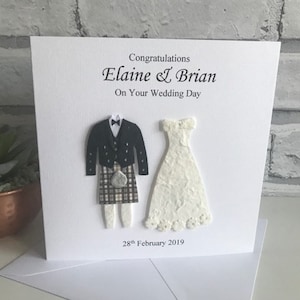 K&ouml;nnte beinhalten: Eine wei&szlig;e Karte mit einer Hochzeitsgl&uuml;ckwunschbotschaft f&uuml;r Elaine & Brian. Die Karte zeigt eine Papierschnittfigur einer Braut in einem wei&szlig;en Kleid und eines Br&auml;utigams in einem Kilt. Das Datum "28. Februar 2019" ist auf der Karte gedruckt.