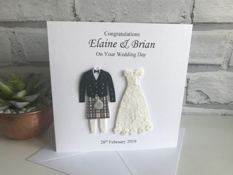 K&ouml;nnte beinhalten: Eine wei&szlig;e Karte mit einer Hochzeitsgl&uuml;ckwunschbotschaft f&uuml;r Elaine & Brian. Die Karte zeigt eine Papierschnittfigur einer Braut in einem wei&szlig;en Kleid und eines Br&auml;utigams in einem Kilt. Das Datum "28. Februar 2019" ist auf der Karte gedruckt.