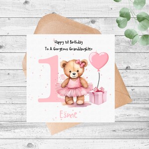 May include: A square greeting card featuring a brown teddy bear wearing a pink tutu and bow, a large pink number one, a pink heart-shaped balloon, and a pink gift box. The text reads Happy 1st Birthday To A Gorgeous Granddaughter and Esme.