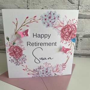 Pu&ograve; includere: Un biglietto bianco con un design a corona di fiori. La corona &egrave; composta da fiori rosa e bianchi, foglie verdi e rami dorati. Il biglietto dice "Happy Retirement Susan" in lettere nere. Ci sono due farfalle rosa con centri bianchi sul biglietto.