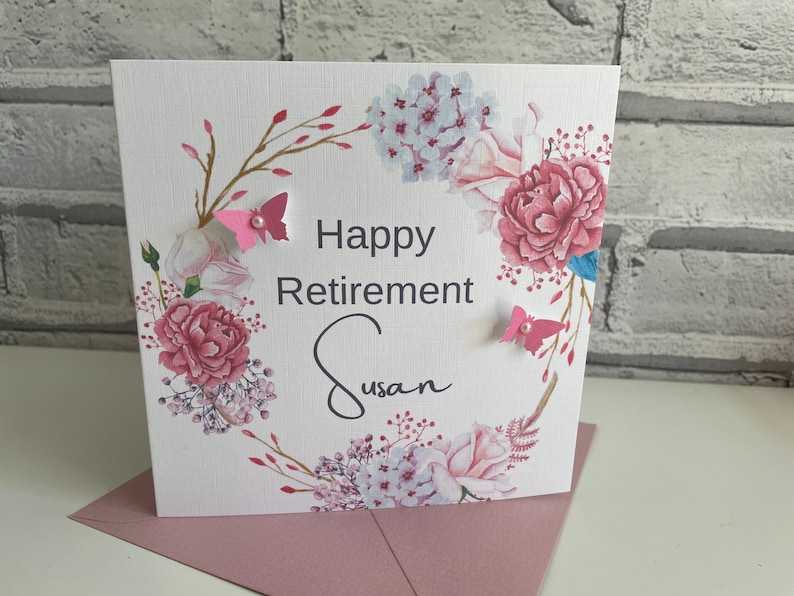 Pu&ograve; includere: Un biglietto bianco con un design a corona di fiori. La corona &egrave; composta da fiori rosa e bianchi, foglie verdi e rami dorati. Il biglietto dice "Happy Retirement Susan" in lettere nere. Ci sono due farfalle rosa con centri bianchi sul biglietto.