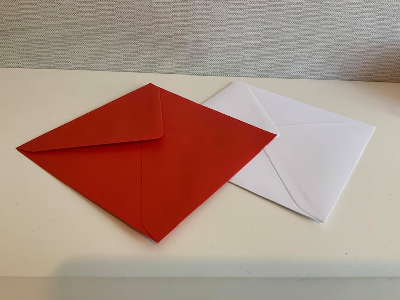 May include: Two envelopes, one red and one white, are lying flat on a white surface. The red envelope is on top of the white envelope.