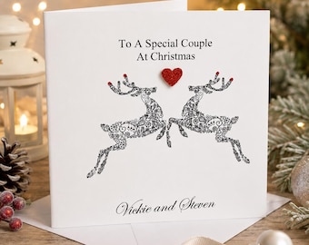 May include: A white square greeting card featuring the black text To A Special Couple At Christmas at the top. Below, two black filigree reindeer face each other with a small red glitter heart between them. The names Vickie and Steven are printed in black script at the bottom.