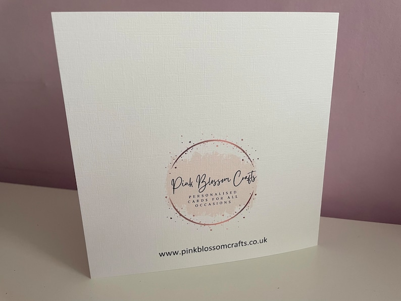 May include: A square white greeting card with a textured surface. The centre features a circular logo with a light pink watercolour wash, rose gold speckles, and the text Pink Blossom Crafts, Personalised Cards For All Occasions. The website www.pinkblossomcrafts.co.uk is printed in black at the bottom.