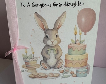 May include: A pink and white birthday card featuring a bunny rabbit, a cake, and a balloon. The card reads "Happy 1st Birthday To A Gorgeous Granddaughter" and "Amber Rose".