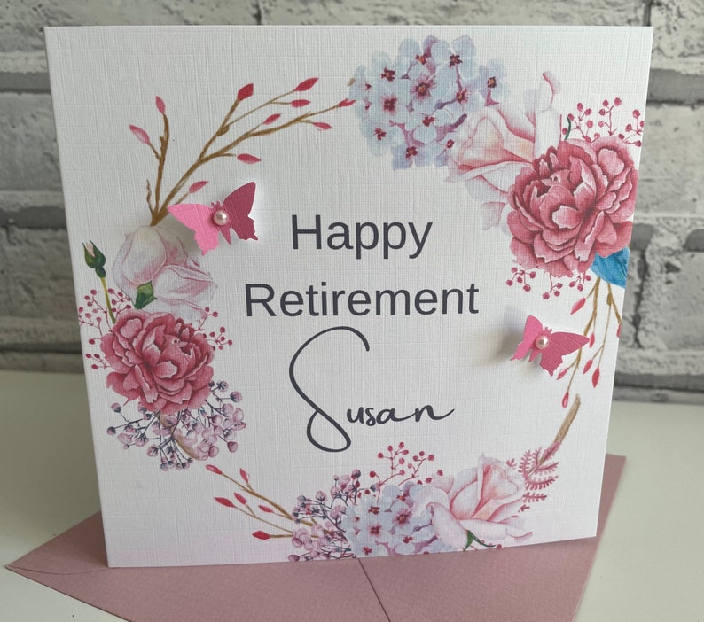 Pu&ograve; includere: Un biglietto bianco con un design a corona di fiori rosa. Il biglietto dice "Happy Retirement Susan" in scrittura nera. Ci sono farfalle rosa con accenti di perle sul biglietto.