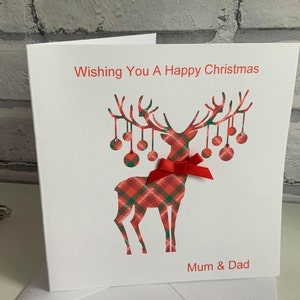 May include: A white greeting card featuring a red and green plaid reindeer silhouette with hanging ornaments on its antlers. A small red ribbon bow is attached to the reindeer's neck. The text reads Wishing You A Happy Christmas and Mum & Dad.