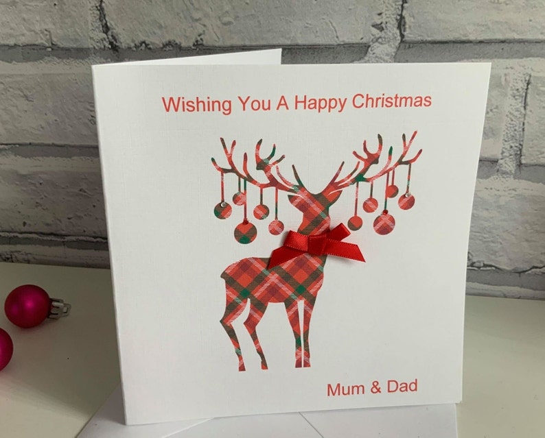May include: A white greeting card featuring a red and green plaid reindeer silhouette with hanging ornaments on its antlers. A small red ribbon bow is attached to the reindeer's neck. The text reads Wishing You A Happy Christmas and Mum & Dad.