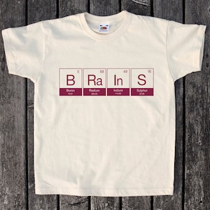 May include: A cream-coloured t-shirt featuring the word "BRAINS" created from periodic table elements: Boron, Radium, Indium, and Sulphur. The text is burgundy. The t-shirt has a crew neck and short sleeves.