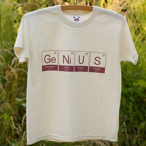 May include: A white t-shirt with a brown print of the periodic table elements spelling out the word "Genius".